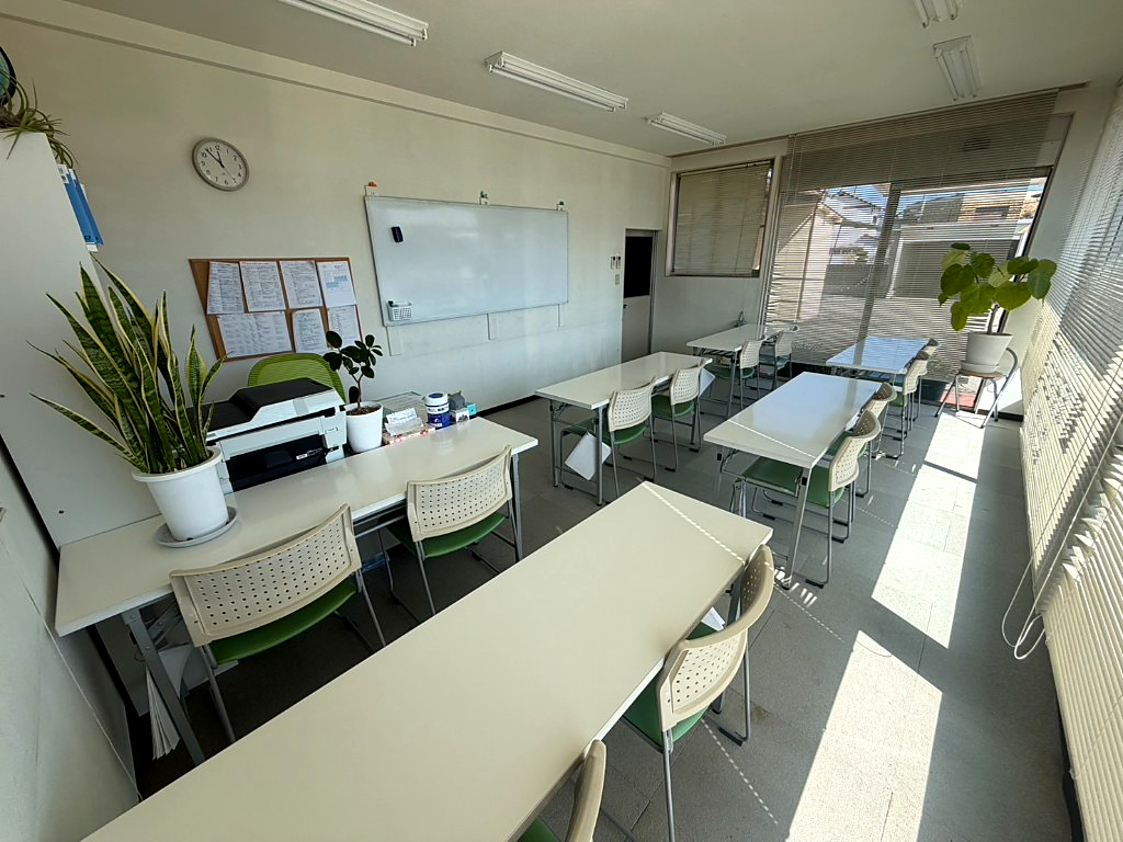 坂東ゼミの教室。自然光の入る明るい空間に白い机・緑のいす、ホワイトボード、掲示板が見える